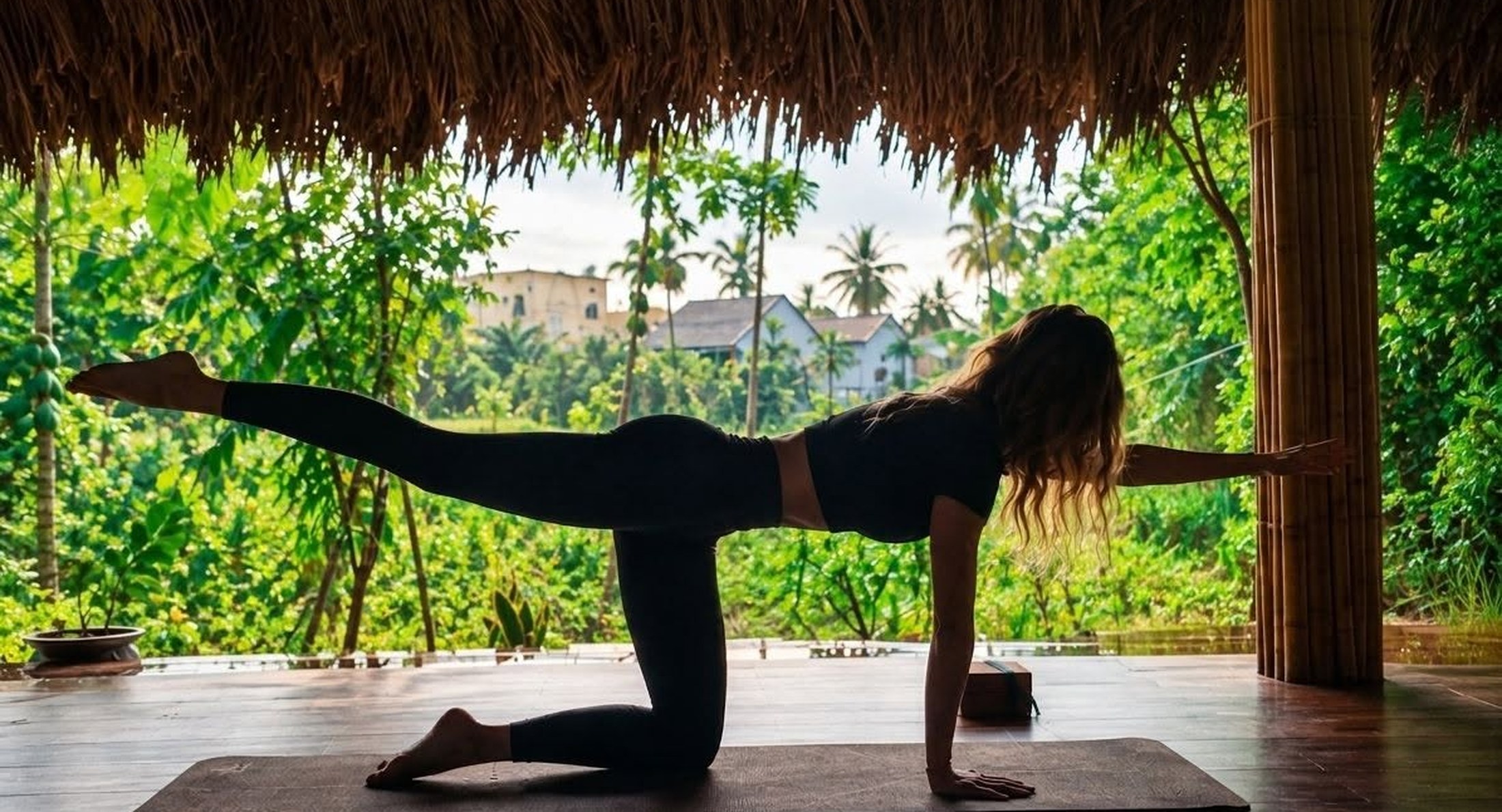 Silhouette of a woman in a yoga balance pose on a mat in an open bamboo studio overlooking green trees